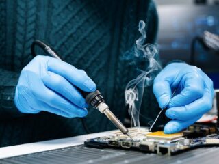 Ilustrační stock: ruka pájecím hrotem nad elektronickou deskou, kouř stoupá vzhůru, civilně bez loga