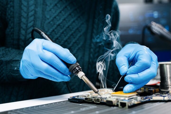 Ilustrační stock: ruka pájecím hrotem nad elektronickou deskou, kouř stoupá vzhůru, civilně bez loga