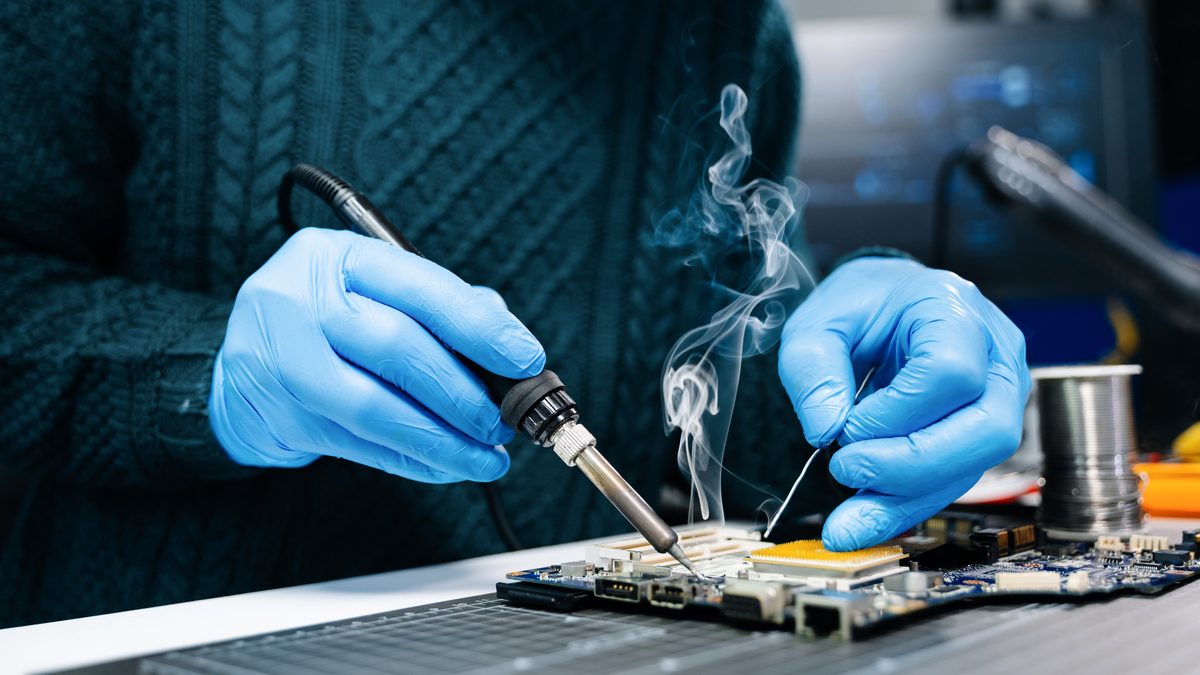 Ilustrační stock: ruka pájecím hrotem nad elektronickou deskou, kouř stoupá vzhůru, civilně bez loga