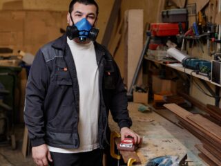 Ilustrační stock: muž v šedé polomasce s výměnnými filtry drží rukou bílý respirátor FFP2, na pozadí rozmazané dílenské prostředí bez loga